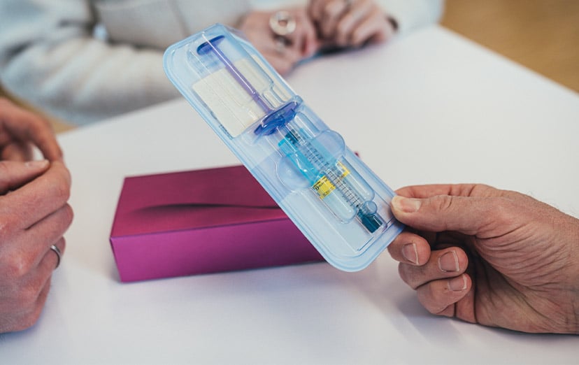 Hand entnimmt eine vorfüllbare Spritze aus einer Blisterverpackung, im Hintergrund eine Produktschachtel